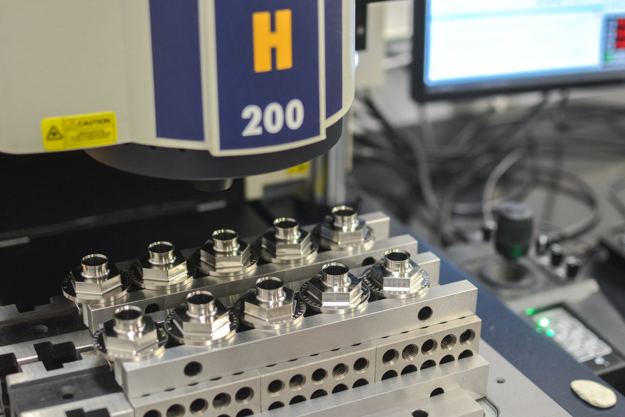 close-up of metal parts in a precision engineering context, positioned under an industrial machine marked "h 200" in an inspection lab.