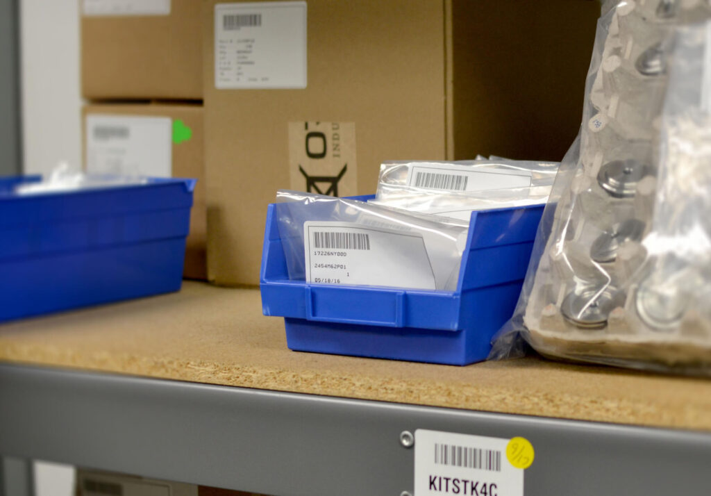 blue storage bins with labels sit on a shelf in a supply room, filled with packaged assembly solutions, alongside larger boxes and clear plastic bags.