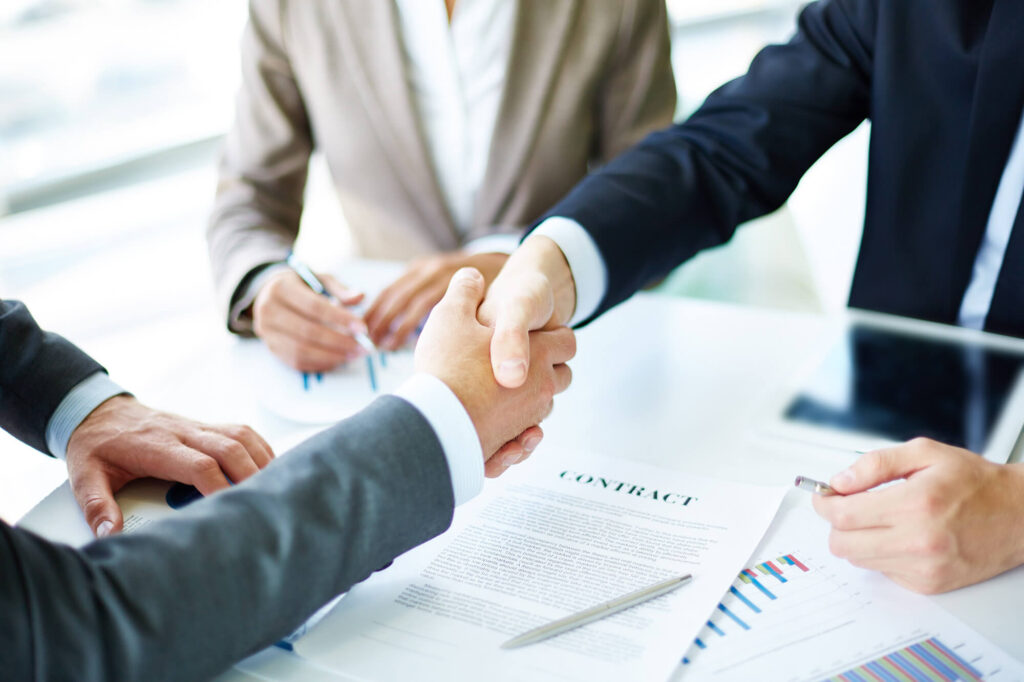 two individuals shaking hands over a desk with a contract, in the presence of others, indicating a business agreement.